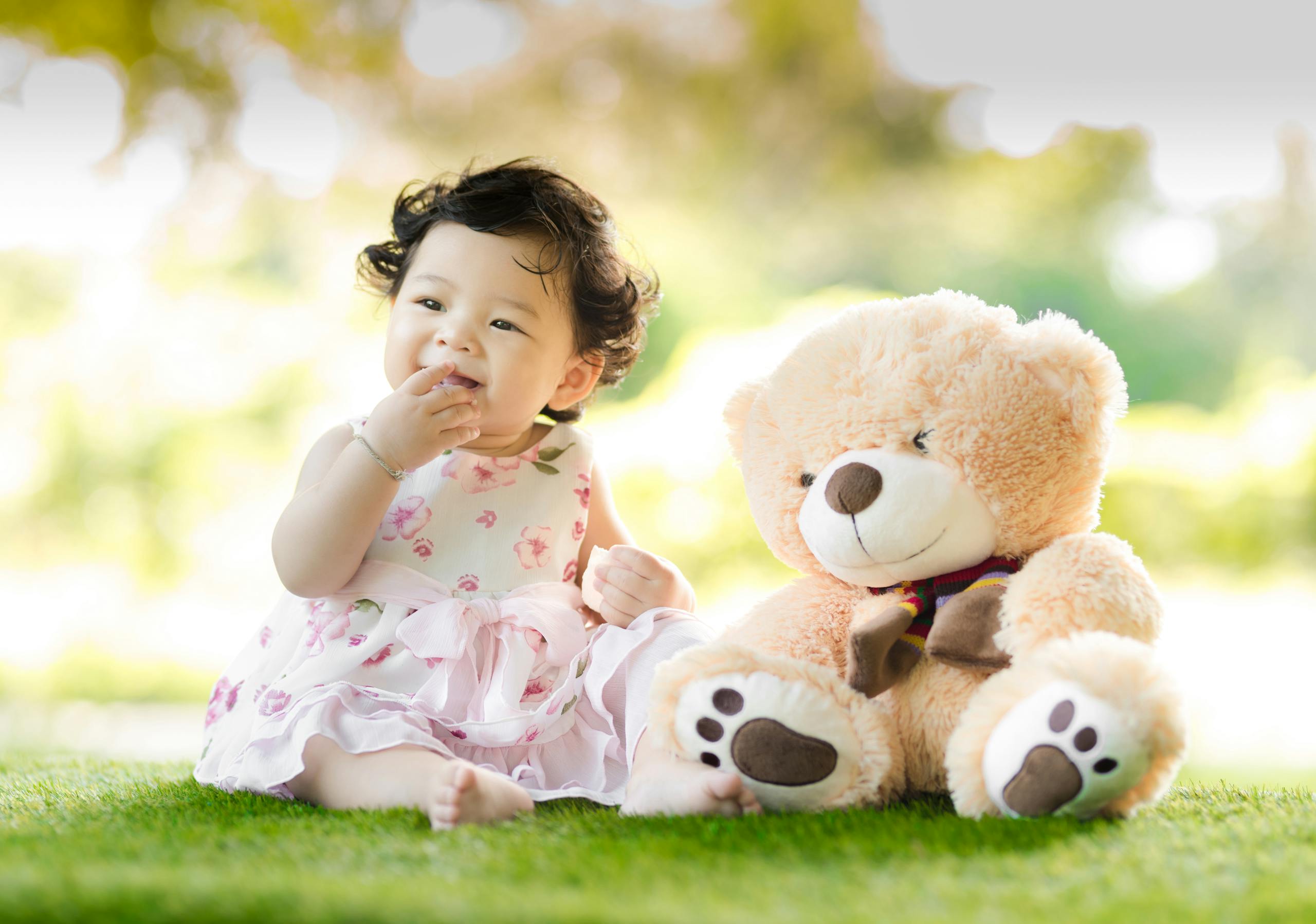 Smiling baby girl sits on grass with a plush teddy bear, enjoying a sunny day.